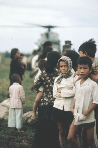 Children standing in a field at the site of a helilift being performed to give aid to survivors of a Viet Cong attack on the village of Vinh Quoi, Vietnam. A boy and girl look directly into the camera in the foreground. Out of focus in the background are other survivors, soldiers and a helicopter.
