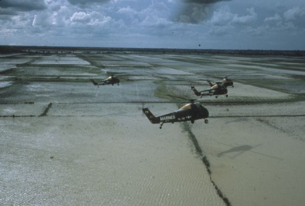 Aerial view of four U.S. Marine helicopters banking over an expanse of rice paddies in Vietnam. Fluffy white clouds fill the sky.