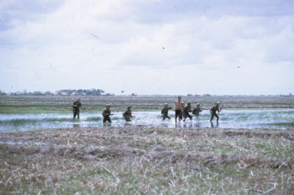 Vietnamese Rangers and U.S. Army Ranger advisor ford a rice paddy with a captured prisoner suspected of being Viet Cong in the vicinity of Bac Lien, Vietnam. The prisoner has his elbows bound behind his back and the Rangers carry rifles. There are buildings in the far background.