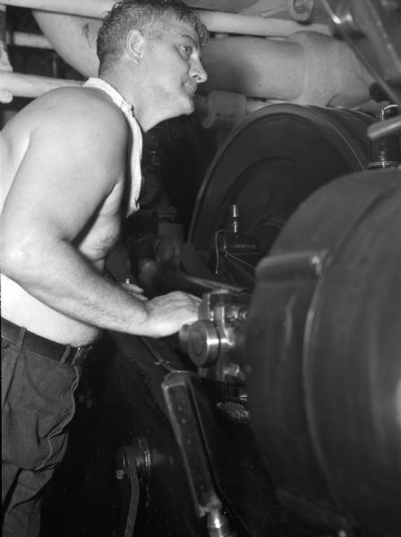 3rd engineer aboard the United Fruit Company's S.S. <i>Santa Marta</i> with a telegraph machine. The engineer is shirtless with a towel around his neck. He is leaning against the large machine. Behind him are pipes. This photograph was taken in the Caribbean.
