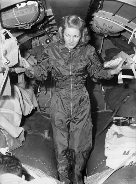 Flight nurse, Gwen Jensen, on a plane with wounded soldiers. Jensen is wearing a jumpsuit. There are stretchers attached to the sides of the plane with wounded in them. Jensen has her arms out to hold onto the stretchers.