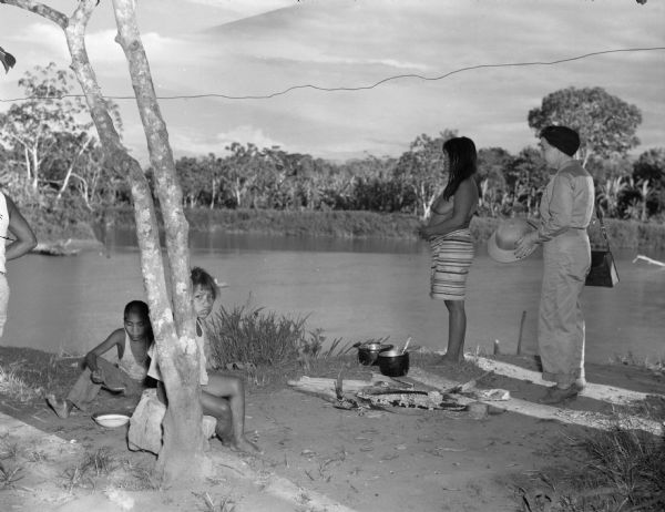 Indigenous Panamanian women and children eating dinner along the Chucunaque River. A woman in a jumpsuit holding a hat is watching Panamanian children eating. A Panamanian woman is standing by two pots and food and the remains of a fire. Behind them is the Chucunaque River and trees.