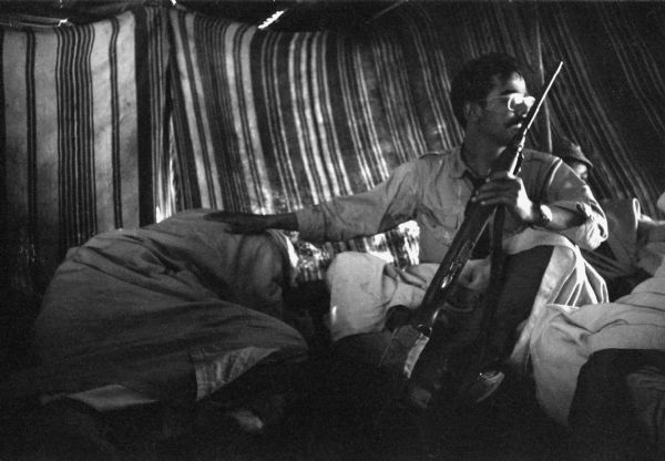 An Algerian member of the National Liberation Front lying down on the ground as another member grabs a rifle and looks out of the tent. He has his hand protectively on the man crouching.