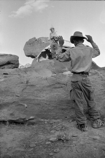 Algerian National Liberation Front member captures an Algerian traitor who helped the French. The FLN member, wearing grass on his hat and shirt as camouflage, is standing high on a boulder pointing a rifle at the traitor. The traitor has his hands raised in surrender in the foreground.