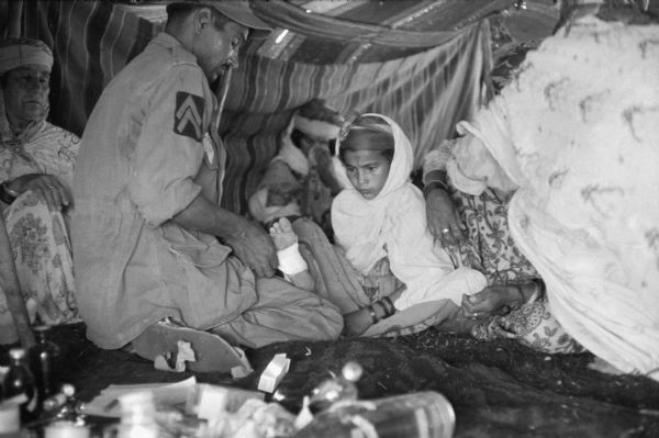 A young Algerian girl has her foot treated by a Red Crescent (Red Cross equivalent) worker in a tent. The man is wrapping the girl's foot in bandages. She is wearing a cloak and a patterned dress. Beside her are two women and behind her is another woman with a baby. There are medicine bottles in the foreground.