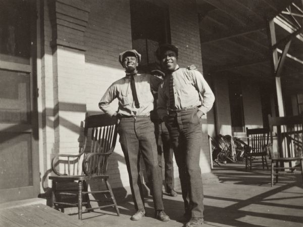 Snapshot of two African American men standing on a porch by a wooden chair in front of a brick building. A third man is standing behind them. The men are wearing hats, button-up shirts and ties.