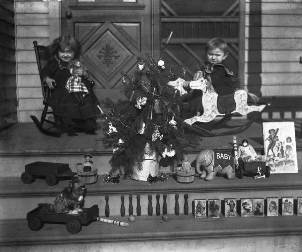 Jennie, in rocking chair, and Edgar Krueger, on a rocking horse, with small Christmas tree and toys, including wagon, blocks, and doll, displayed outside on the steps of the porch.