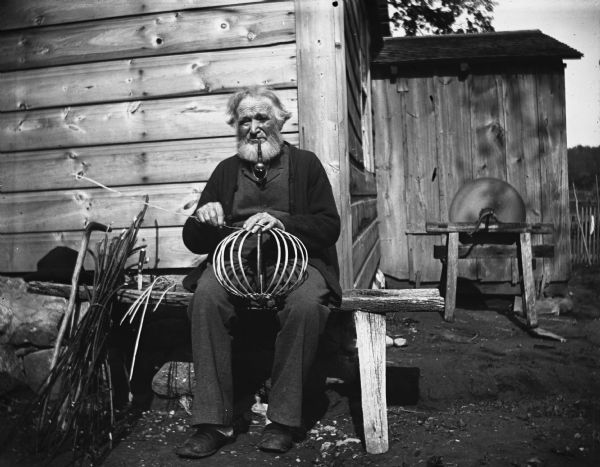 View of William Krueger sitting on a bench next to the house, weaving a willow basket and smoking a pipe. In the background is a pedal grinding/sharpening wheel.
