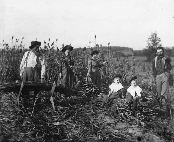 August, Tina, Sarah, Jennie, Edgar and Alex Krueger posing outdoors in a field. Alex took this photograph remotely with a string tied to the shutter of the camera. They are all wearing hats except Alex, and the two young children have large collars over their clothing.