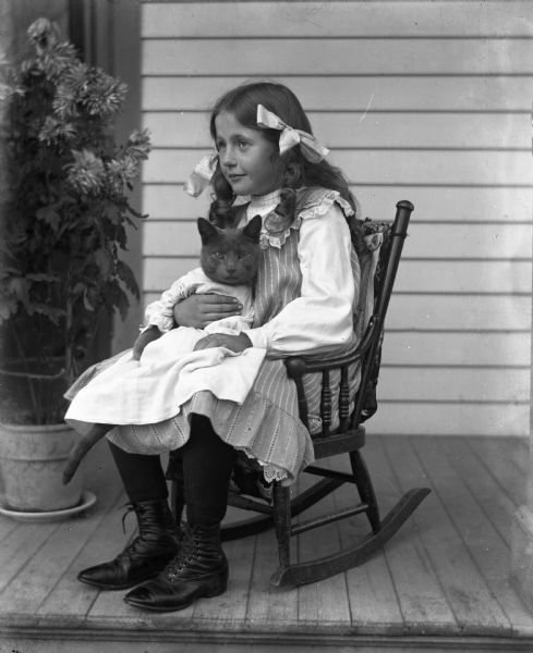 Jennie Krueger sitting in a rocking chair on a porch holding her cat Tramp, who is dressed in baby clothes.