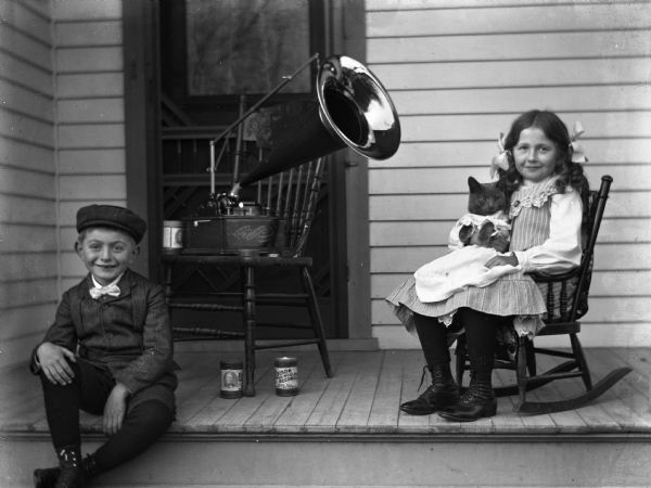 Edgar and Jennie Krueger are sitting on the front porch of the Krueger home with an Edison Standard phonograph sitting on a chair between them. Two Edison Gold Moulded Records cylinders are under the chair. Jennie is holding their cat named "Tramp" on her lap. The cat is wearing a dress.