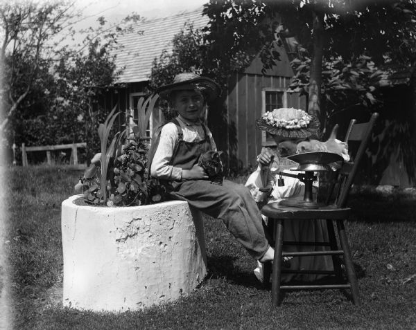 Edgar and Jennie Krueger outdoors in the yard. Edgar is sitting on a concrete planter holding a kitten in his lap. Jennie is kneeling in the grass weighing kittens on a scale set up on a chair.