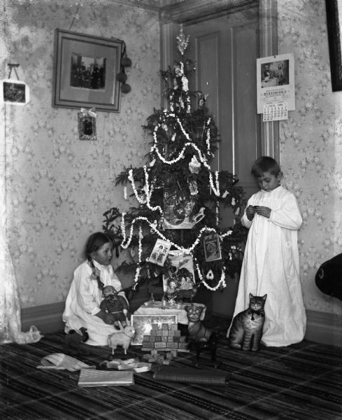 Young children, Jennie and Edgar Krueger, playing with toys near their family Christmas tree.