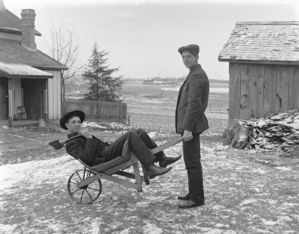 Henry Bigalk relaxes in a wheelbarrow holding an axe in his left hand, while Emil Kuney (Keune) is grabbing the wheelbarrow handles as if to transport Bigalk away. Behind them is an outbuilding on the right, and a house is on the left. In the far distance across a field are more buildings. Snow is on the ground.