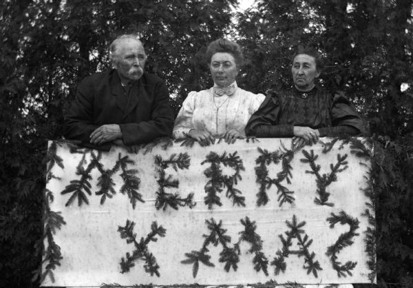 August Krueger Family Christmas August, Sarah, and Mary Krueger supporting a Merry Xmas sign made from pine branches.