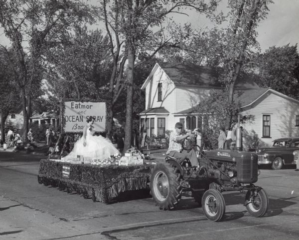 Parade float sponsored by Eatmor and Ocean Spray Cranberries. A beauty queen sits on the float surrounded by cranberry products. The float is being pulled by an International Harvester Farmall A tractor.