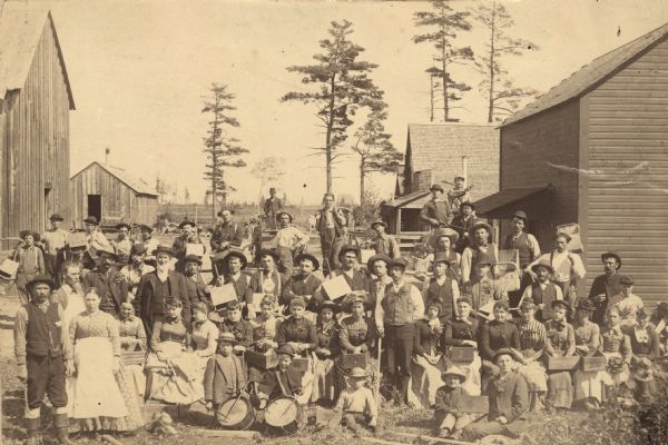 Large group of cranberry harvesters posing with implements. Many of the individuals are wearing boutonnières and corsages made from cranberry sprigs. Many of the implements have the initials C&S printed on them.