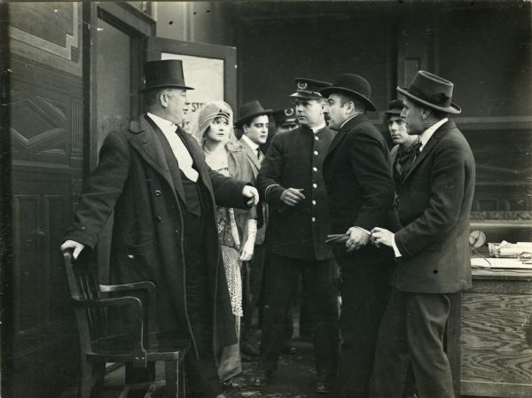 Stirling, the corrupt Wall Street broker, is confronted by Mr. Berry (played by W. H. Brown in a top hat) in the climax of the 1915 Reliance one-reel silent film "Above Par." Behind Berry is his daughter (Billie West) who is the fiancée of the newspaper reporter Dick Carson (W. E. Lawrence, behind West in the light-colored suit). The police officer is probably played by Jack Dillon.