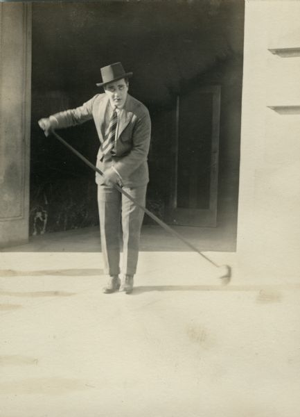 A publicity still of the actor W.E. Lawrence cleaning up with a push broom, or at least pretending to do so.
