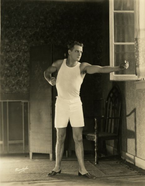 Pat Glendon, Jr. (played by Reginald Denny) strikes a boxing pose near an open window. He wears white athletic shorts, an undershirt, and house slippers. An contemporary caption written on the back of the print notes appreciatively, "'The Abysmal Brute' has the physique of a young god."
