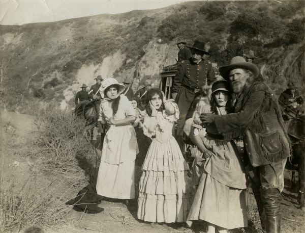 Three or more pioneer women and an older man in buckskin look in horror at a threat--indians?--that is beyond the frame of the photograph in this scene still from the one-reel silent film "Across the Plains," also known as "War on the Plains." The young woman in the center is the actress Ethel Grandin. Behind the group is a stagecoach and a U.S. Army calvary escort.