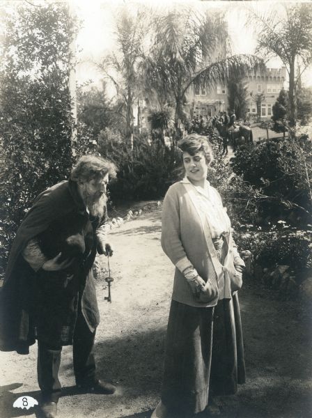 A stooped old man with a long beard approaches Kitty Gray (played by Grace Cunard) with some large keys in a scene still from Episode 8 "The Prison in the Palace" of the 1915 Universal serial "The Broken Coin." They are in a garden, and far behind them is a palace and a group of mounted soldiers.