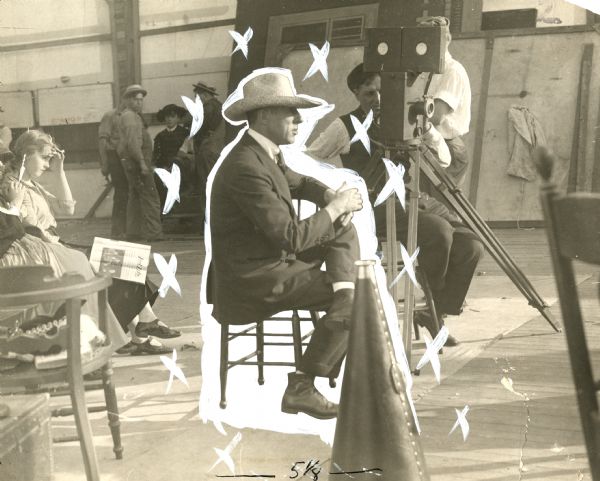 Director D.W. Griffith wears a straw hat and sits watching a scene being filmed in a production still that has been heavily retouched for print reproduction. Behind him is a Pathé motion picture camera which obscures cameraman G.W. "Billy" Bitzer and his assistant Karl Brown. On the left, the young actress Dorothy Gish sits watching the scene with a copy of "Reel Life" magazine on her knee.