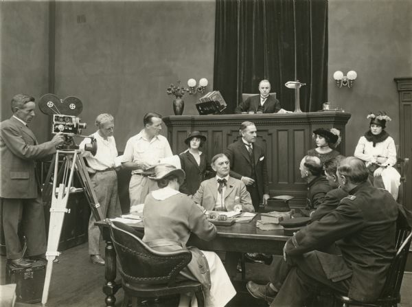 Original caption: "Director Phillips Smalley (third from the left), rehearsing his players in Brand Whitlock's story being filmed at Universal City. The judge is Roy Stewart, and the lawyer standing in front is Dana Ong, former Assistant District Attorney of Los Angeles. The two girls at the right are Irene Aldwyn and Hazel Page, who play the leads with Stewart."