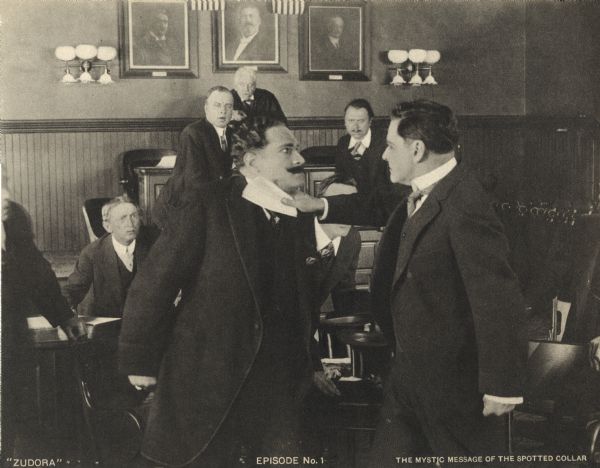 John Storm (played by Harry Benham) slaps a moustached man (Arthur Bauer) with a piece of paper while a judge, witness (John Reinhard, rear right), court clerk (N.Z. Wood, half-seated at left), and others look on in a scene still from the Thanhouser serial "Zudora." The print is captioned "Episode No. 1" and "The Mystic Message of the Spotted Collar."