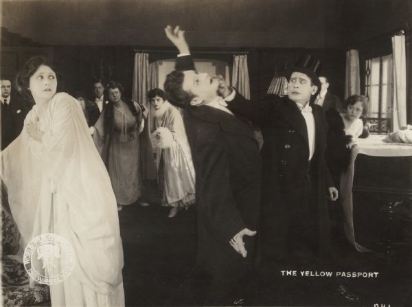 Sonia Sokoloff (played by Clara Kimball Young in a flowing white gown) shrinks back in alarm as Fedia (John St. Polis) receives a terrific right punch to the jaw from Adolph Rosenheimer (Edwin August). In the background other well-dressed men and women react to the fight.