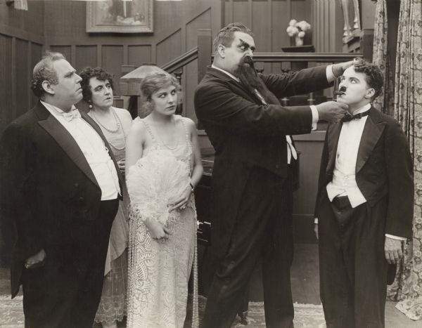 In this scene still from Chaplin's 1917 silent film "The Adventurer," Eric Campbell, made-up with large eyebrows, beard, and moustache, closely scrutinizes Charlie Chaplin's face. Watching, from left to right, are Henry Bergman, Marta Golden, and Edna Purviance.