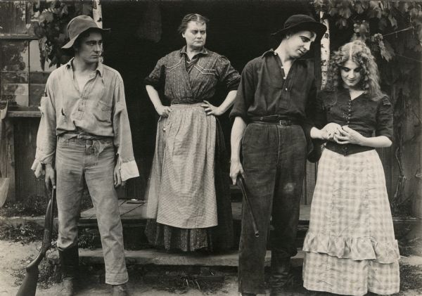 Margie (played by Florence Lawrence, far right) is loved by both Rob (Owen Moore, on the left) and Orin (Gladden James, right) in this publicity still from the 1912 Victor production "After All." Margie's widowed mother (played by Victory Bateman) stands on the stoop. All the actors are costumed in rustic clothes, and the two men carry single barrel shotguns.