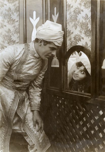 Jack Stanton and Faimeh Fitzgerald (played by Antonio Moreno and Edith Storey) flirt through a screen in a publicity still for "Aladdin from Broadway." They are in Turkish costumes with Moreno wearing a turban and Storey a headscarf.
