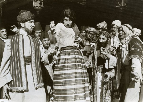 Amad (played by Otto Lederer in beard and turban) watches as his wife Faimeh (Edith Storey) threatens to do herself harm with a dagger in a scene still for "Aladdin from Broadway." They are in Turkish costume and are surrounded by a crowd of actors wearing turbans, fezzes, and one man in an Arab keffiyeh and akal.
