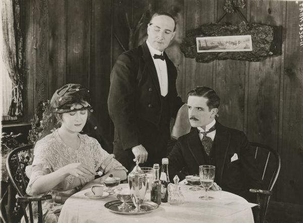 Cicely Osborne (played by Mary MacLaren) adds a sugar cube to her coffee as Philip Ashton (Stanhope Wheaton) who is seated next to her and a waiter watch closely in a scene still for "The Amazing Wife." They are seated at a table in a rustic cafe.
