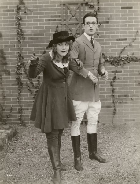 Mildred Davis points off screen while Harold Lloyd looks alarmed. Both wear riding clothes in a scene still for "Among Those Present."