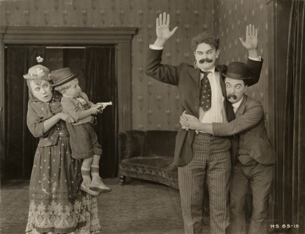 Louise Fazenda holds baby Don Marion who holds a revolver on Eddie Gribbon who is held about the waist by Billy Bevan in a scene still for the Mack Sennett comedy "Astray from the Steerage" (1921). Fazenda, Marion, and Bevan are costumed as recent immigrants. Bevan and Gribbon both wear exaggerated silent comedy moustaches and eyebrows.