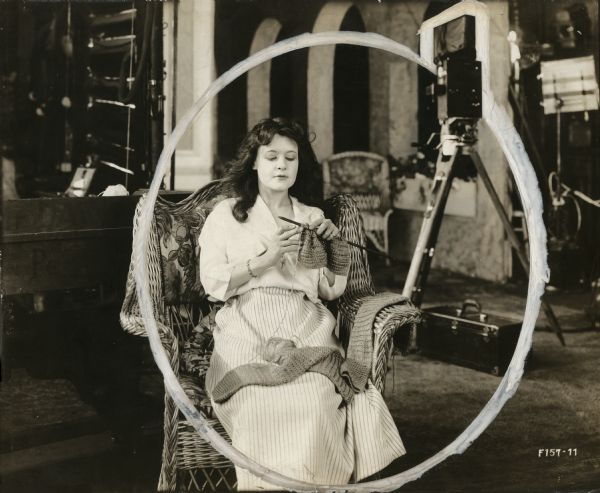Silent film actress Marguerite Clark sits in a wicker chair and knits on a set for "Bab's Burglar." Behind her is a Pathé 35mm motion picture camera on a tripod.