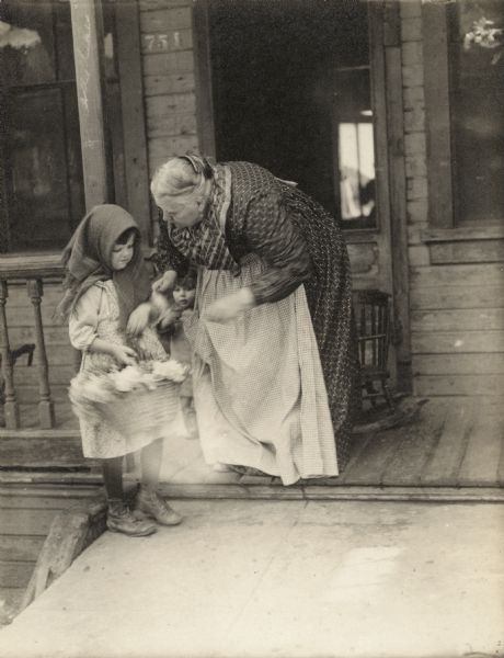 Actress Jennie Lee bends over a small flower girl who wears a kerchief on her head. They stand on the front porch of an impoverished house in this scene still from the short silent film "The Balance" (1915).