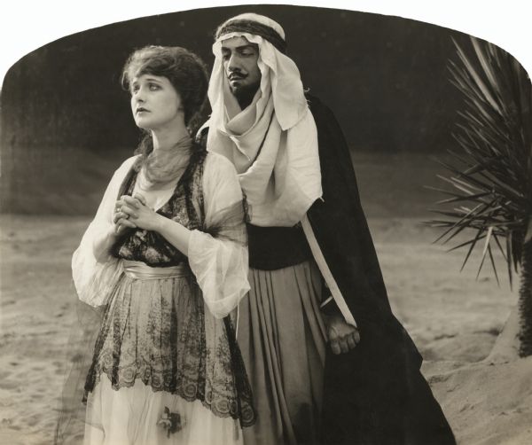English noblewoman Kathryn, Lady Wyverne (played by Elsie Ferguson), stands in a prayerful attitude in a scene still for the 1917 silent film "Barbary Sheep." She wears a white dress with a black lace coverlet. Looming close behind her is the Arab chieftan Benchaalal (played by Pedro de Cordoba). He wears a keffiyeh and dark robes and has a Van Dyke beard.