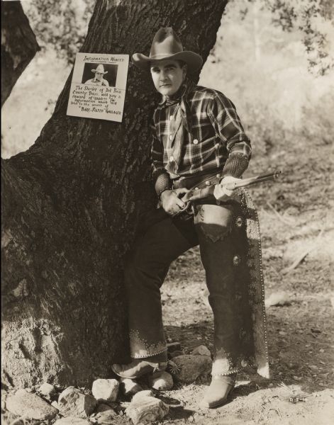 Bare-fisted Gallagher (played by William Desmond) stands in full Western costume (cowboy hat, plaid shirt, bandana neckerchief, charro chaps, leather wrist cuffs) looking watchful with a Winchester carbine repeating rife in his hands. A wanted poster on the tree behind him says that the sheriff is offering a thousand dollar reward for him.