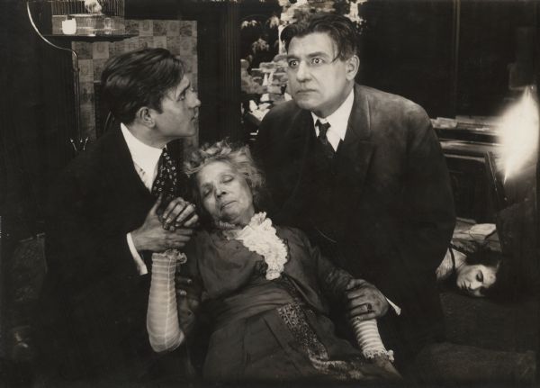 In a dramatic scene still from the silent film "The Battle Cry of Peace," Charley Harrison (left, played by James Morrison) and John Harrison (right, played by Charles Richman) support their mother (Mary Maurice) who has fainted. New York City is being invaded by the enemy, and Virginia Vandergriff (played by Norma Talmadge on the floor, lower right) has been shot by her own mother to prevent her violation by the enemy.