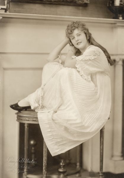 In this Campbell Studio publicity portrait, the 23 year old actress Grace Darling sits on a small table and wears a white, pleated gown. The portrait was used to publicize the Wharton silent film serial "Beatrice Fairfax" in which Darling played the title character, a newspaper advice columnist and crime fighter.