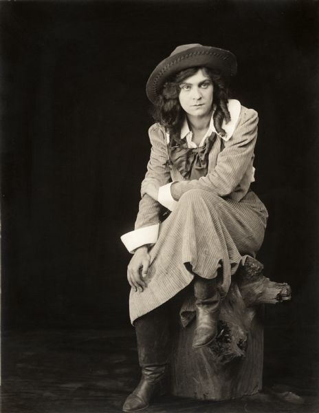 In a publicity still for the Lubin silent film series "The Beloved Adventurer," Lottie Briscoe wears a corduroy western dress, boots, and a cowboy hat.