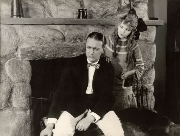 Actress Gladys Leslie puts a hand on Huntley Gordon's shoulder as he sits by a rustic fireplace looking worried in a scene still for the 1918 Vitagraph drama "The Beloved Imposter."