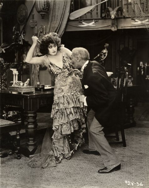 Lolette, the Spanish peasant girl (played by Theda Bara) uses a knife to defend herself from a middle aged man in a scene still for the silent drama "The She-Devil." She wears a Spanish dancing costumes with many ruffles. The room is exotically decorated with knives, a mounted ram's head, a boat paddle, a fishing net, a Chinese lantern, and so on.