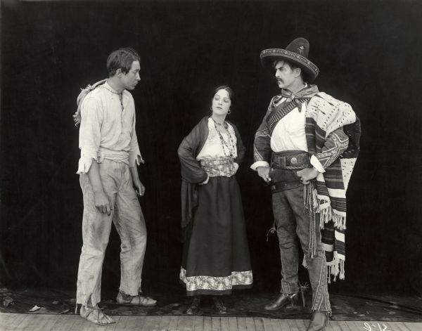 In this publicity still for "Betrayed," a 1917 Fox silent drama set in Mexico, the actors Monte Blue, Miriam Cooper, and Hobart Bosworth pose in costume against a black background. Bosworth plays the bandit Leopoldo Juares whose costume is a particularly resplendent example of vaquero dress with sombrero, spurs, wide belt, bandolier, sarape, and so on.
