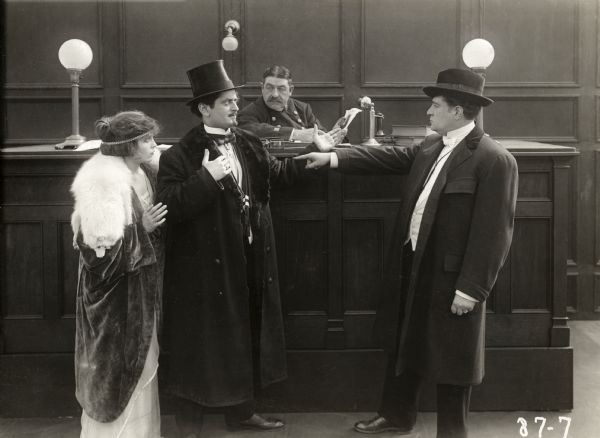 In a scene still set in a police station for the 1915 Thanhouser silent drama "Bianca Forgets" are Bianca Wells (played by Florence La Badie wearing a white fox stole), Count Berdeau (played by Charles A. Jahn), and a police detective (played by Arthur Bauer). Behind them is the desk sergeant, played by an unidentified actor.