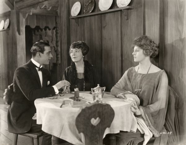 Lord Hector Bracondale (played by Rudolph Valentino) has a heart to heart talk with his mother Lady Bracondale (Edythe Chapman) as Morella Winmarleigh (Gertrude Astor) looks on coldly. This scene still for the silent drama "Beyond the Rocks" is set in a rustic dining room.