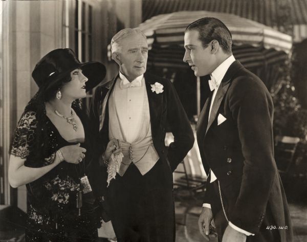 Theodora Fitzgerald and her father Captain Fitzgerald (played by Gloria Swanson and Alec B. Francis) talk to Lord Hector Bracondale (Rudolf Valentino) in a scene still for the silent drama "Beyond the Rocks." All are in formal evening wear.
